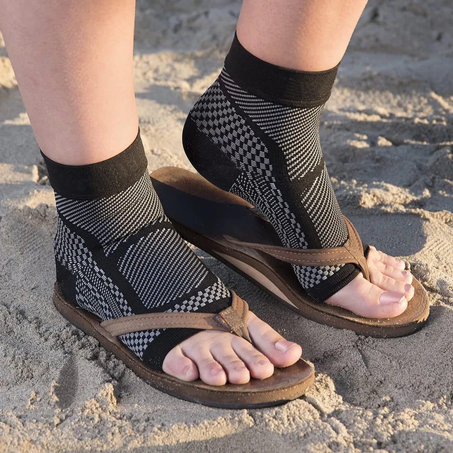 Feet wearing black compression socks with sandals on sand, showing arch support.