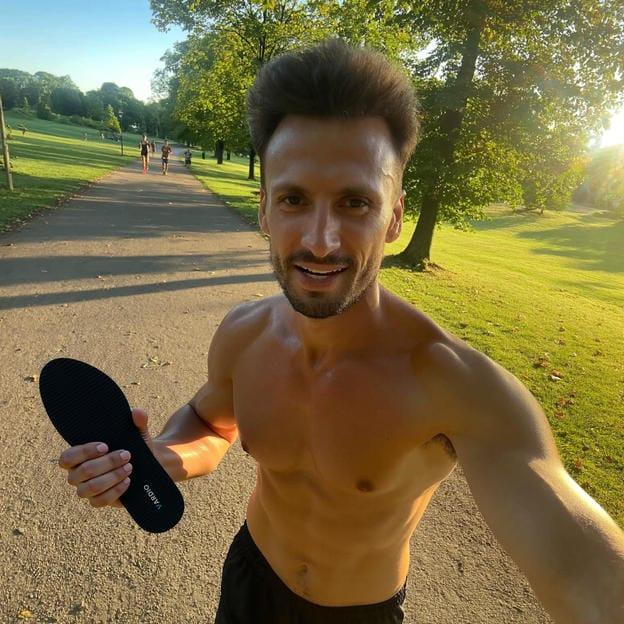 Shirtless man outdoors holding black insole on running path in sunny park
